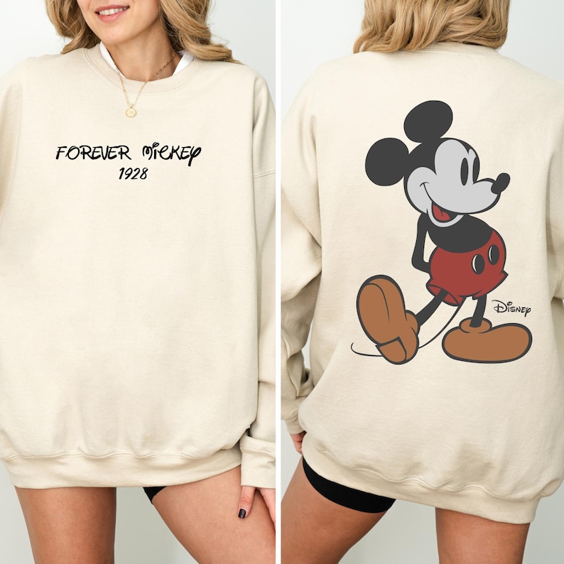 Puede incluir: Sudadera color crema con el texto "FOREVER MICKEY 1928" en escritura negra en la parte delantera. La parte trasera muestra un gran gr&aacute;fico de Mickey Mouse en su pose cl&aacute;sica, con el logo de Disney.