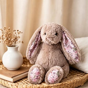 May include: A plush bunny rabbit with long, floppy ears and a soft, textured body. The inside of the ears and the soles of the feet feature a floral pattern in shades of pink and purple. The bunny is sitting on a woven tray.