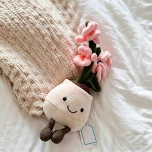 May include: A soft toy plant pot with a smiling face, brown feet, and pink flowers on green stems. It rests on white sheets next to a cream-coloured knitted blanket. The toy is designed to look like a potted plant.