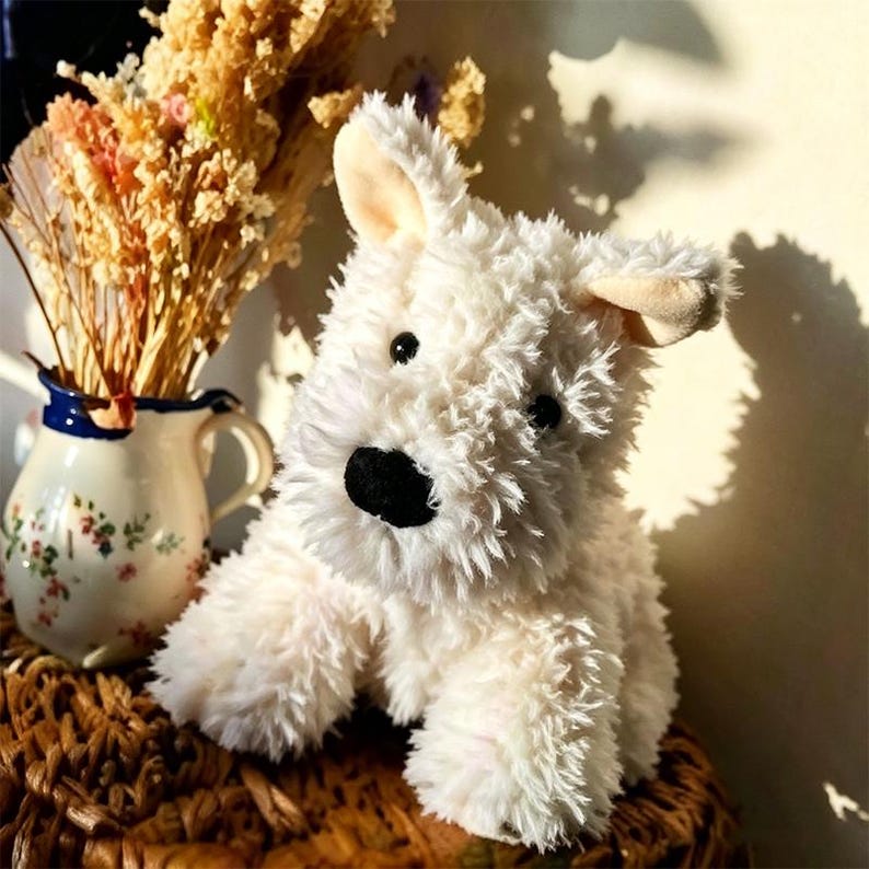 Puede incluir: Un peluche blanco con forma de perro, con nariz y ojos negros. El perro de peluche tiene orejas ca&iacute;das y una textura esponjosa. Est&aacute; colocado sobre una superficie tejida, con una jarra decorativa y flores secas en el fondo.