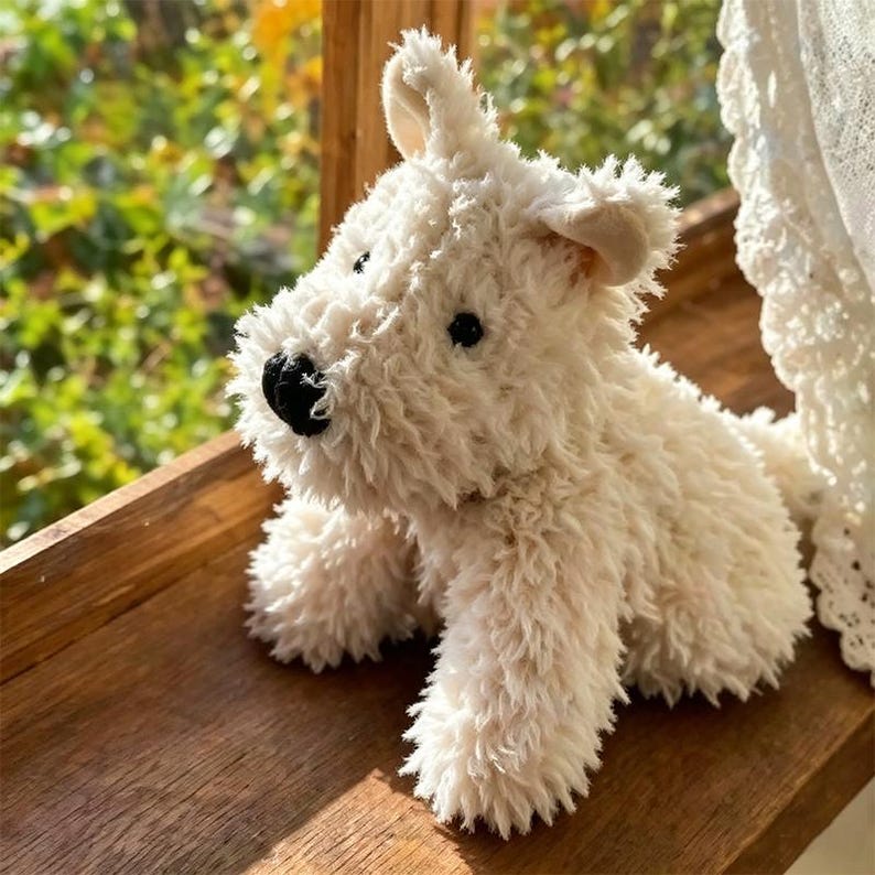 Puede incluir: Un peluche de perro terrier blanco roto con una textura esponjosa. El juguete tiene ojos negros de bot&oacute;n y una nariz negra. Est&aacute; colocado sobre una superficie de madera, con un fondo verde borroso.