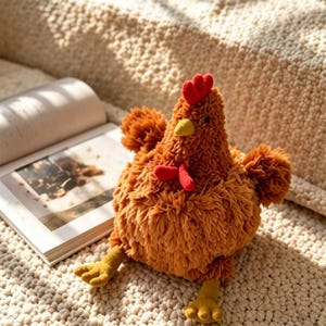 May include: A soft, brown chicken toy with a red comb and wattle, yellow beak and feet. The plush toy is positioned on a cream-coloured surface beside an open book. The chicken has fluffy, textured feathers.