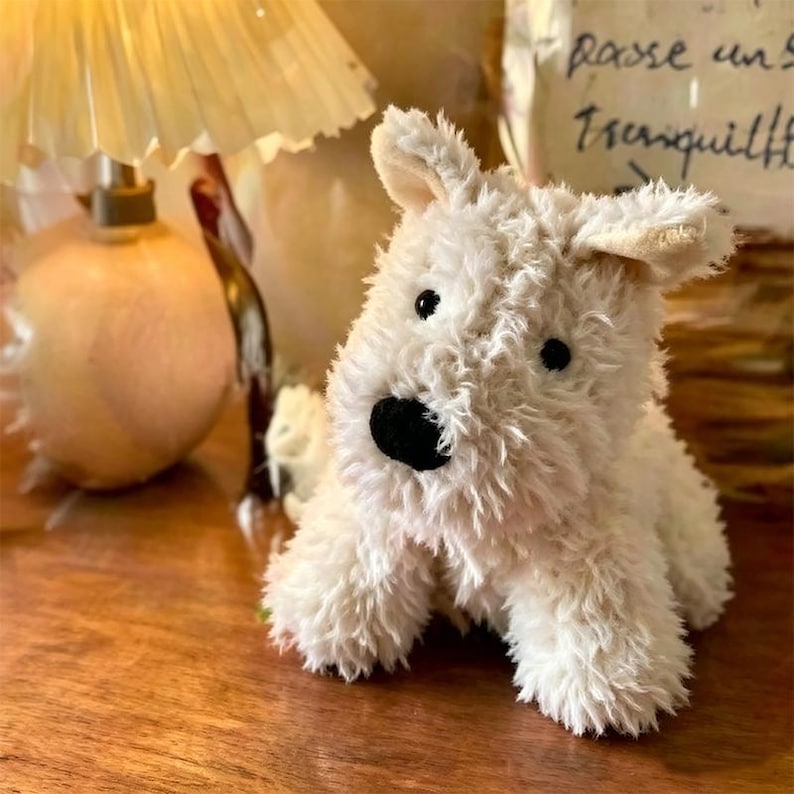 Puede incluir: Un juguete de peluche blanco con forma de perro, de textura suave, ojos negros y nariz negra. El juguete est&aacute; sobre una superficie de madera. Un art&iacute;culo lindo y tierno.