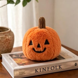 May include: An orange plush pumpkin with a carved face, including triangular eyes and a smiling mouth. The pumpkin has a brown stem and sits atop a book with the word "KINFOLK" on the cover. A decorative Halloween item.