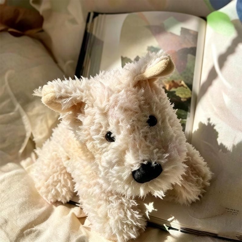 Puede incluir: Un peluche de perro de color crema, con una textura suave. El juguete tiene ojos negros y una nariz negra. El perro est&aacute; tumbado sobre una superficie suave y clara, con un libro abierto en el fondo.