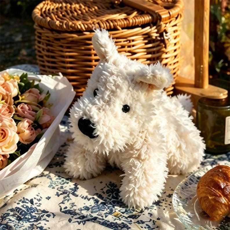 Puede incluir: Un juguete de peluche en forma de perro, de color crema, con nariz y ojos negros. El juguete est&aacute; colocado sobre un mantel estampado, con una cesta de picnic y un ramo de flores en el fondo. Un cruas&aacute;n est&aacute; sobre un plato de cristal.
