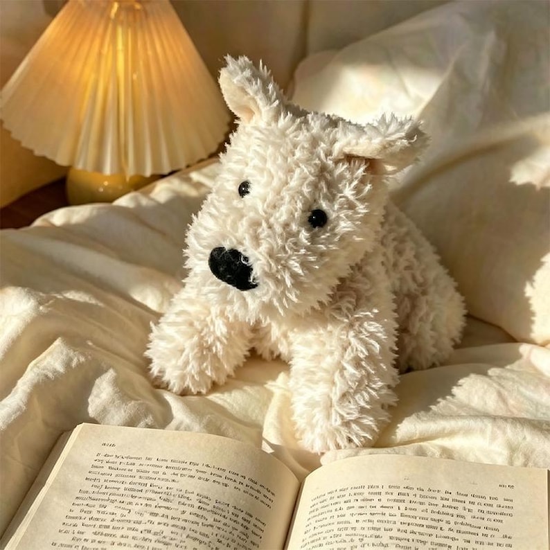 Puede incluir: Un juguete de peluche en forma de perro, de color crema, con nariz y ojos negros, descansa sobre una cama con un libro. El juguete tiene una textura peluda y est&aacute; colocado frente a un libro abierto. Una l&aacute;mpara con pantalla plisada est&aacute; en el fondo.