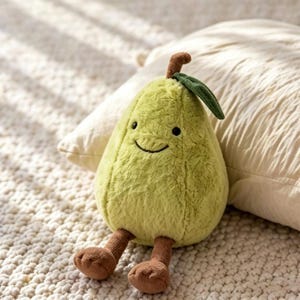May include: A soft, green pear-shaped toy with a smiling face, brown feet, and a stem with a leaf. The plush toy is sitting on a white, textured surface, with a pillow in the background.