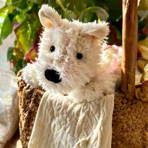 Puede incluir: Un juguete de peluche blanco con ojos y nariz negros, metido en una cesta tejida con una manta de punto de cable color crema. El perro tiene un pelaje suave y orejas ca&iacute;das. La cesta es marr&oacute;n y tiene un asa de madera.