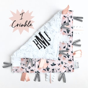 May include: A white and pink baby blanket with a crinkle texture and a variety of ribbons and tags. The blanket is personalized with the initials "HMJ".