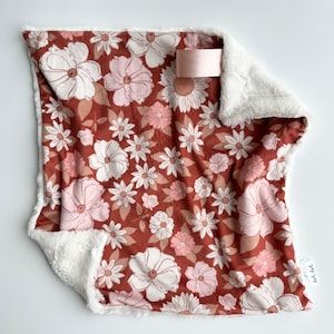 May include: A soft, brown and white floral print baby blanket with a white faux fur border. The blanket has a pink satin tag with the text "didi dot" on it.