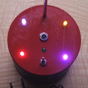 May include: A red, circular electronic device with a black base, featuring a silver antenna and multiple colored LED lights. It has a power adapter with a black cord and plug. The device has a switch and a power input.