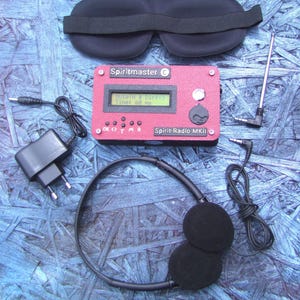 Może przedstawiać: Czerwone urządzenie Spirit-Radio MKII z małym ekranem, przyciskami i napisem "Spiritmaster". Zawiera czarną maskę do spania, słuchawki, antenę i zasilacz. Przedmioty są ułożone na niebieskiej powierzchni.