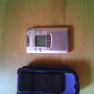 Könnte beinhalten: Ein silberner Panasonic RR-QR240 Digital-Sprachrekorder mit kleinem LCD-Bildschirm, Lautsprechergitter und Bedientasten, begleitet von einer blauen Schutzhülle mit einem schwarzen Stoffriemen auf der Vorderseite.