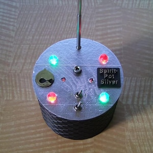 Op de afbeelding: Een zilverkleurig elektronisch apparaat met een cilindrische basis en een cirkelvormige bovenkant. Het heeft meerdere gekleurde LED-lampjes, twee tuimelschakelaars en een kleine antenne. De tekst "Spirit-Pot Silver" staat op een zwarte rechthoekige plaat.