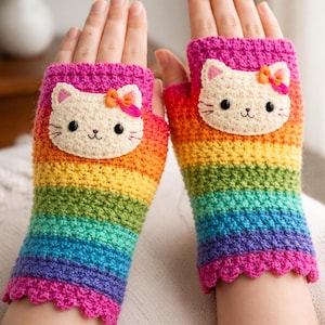 May include: A pair of rainbow-striped, fingerless crochet gloves. Each glove features a cream-colored cat face with black eyes, whiskers, and a pink bow. The cuffs are a bright pink with a scalloped edge, and the stripes transition from red to purple.