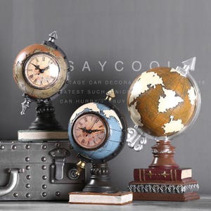 May include: Three decorative globe clocks with vintage styling. One clock is brown and green, another is blue and brown, and the third is a globe with a clear arrow. The clocks have Roman numeral faces and sit on decorative bases.