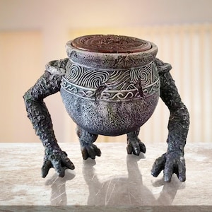 May include: A detailed sculpture of a pot with arms and legs. The grey pot features carved designs and a brown lid. The arms and legs are textured and dark green, giving the sculpture a unique, fantasy-inspired appearance. A decorative item.