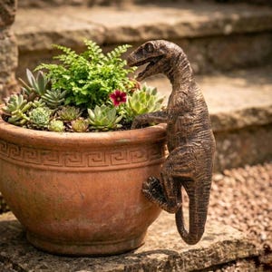 Op de afbeelding: Een terracotta ronde plantenbak gevuld met vetplanten en groen, met een bronzen dinosaurussculptuur die aan de zijkant hangt. De plantenbak heeft een Grieks sleutelontwerp rond de rand. Een kleine rode bloem is zichtbaar.
