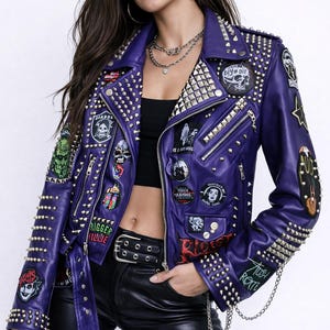 Pode incluir: Uma jaqueta de couro roxa com tachas e patches prateados. A jaqueta tem um estilo motociclista clássico com gola entalhada, zíper assimétrico e vários bolsos. Patches com caveiras e outros gráficos são costurados na jaqueta.