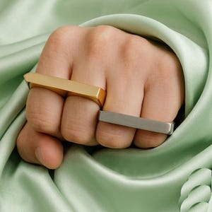May include: A hand displaying two geometric rings. One ring is gold-coloured and rectangular with a pointed end. The other ring is silver-coloured and rectangular. The rings are set against a light green, silky background.