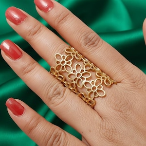 Puede incluir: Un anillo dorado con un diseño floral. El anillo está hecho de una serie de flores caladas, creando un patrón delicado e intrincado. El anillo se lleva en un dedo con esmalte de uñas rojo, sobre un fondo verde.
