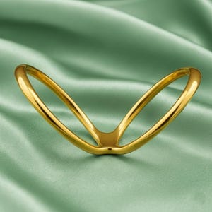 Puede incluir: Un anillo dorado en forma de V con un acabado liso y pulido. El anillo se encuentra sobre un fondo de tela verde suave, creando un contraste de texturas y colores. El diseño es simple pero elegante.