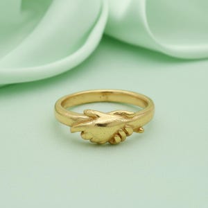 May include: A gold-colored ring featuring a detailed design of two hands clasped in a handshake. The ring is set against a soft, light green background, highlighting the intricate details of the hands.