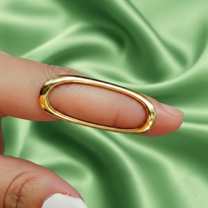 May include: A gold-colored ring with an oval shape. The ring is worn on a finger, and the background is a green, silky fabric.