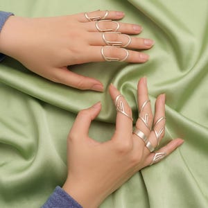 Può includere: Anelli d'argento in vari design adornano le dita, presentando una collezione di gioielli unici. Gli anelli presentano forme geometriche e design aperti, che riflettono un'estetica moderna. Gli anelli sono esposti su uno sfondo di tessuto verde.