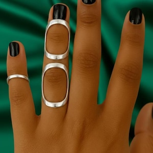 Pode incluir: Uma mão com verniz preto exibe três anéis de prata. Um anel é uma faixa simples, enquanto os outros dois são formas ovais alongadas e conectadas que cobrem dois dedos. O fundo é um tecido verde escuro.