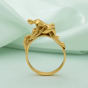 Puede incluir: Un anillo dorado con una figura esculpida en una pose reclinada. El diseño del anillo incorpora la figura como elemento central, con intrincados detalles en el trabajo del metal. El anillo está sobre un fondo verde claro y suave.