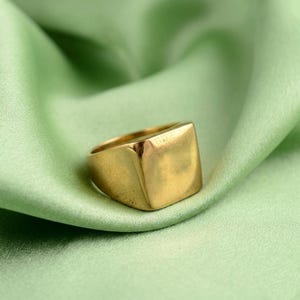 May include: A gold-colored ring with a square face. The ring is set against a backdrop of a light green, silky fabric. The ring's design is simple and modern, with a polished finish.