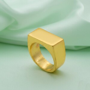 Peut inclure: Une bague chevalière dorée avec un dessus rectangulaire. La bague a une finition lisse et polie et une bande épaisse. La bague est présentée sur un fond vert clair et doux.