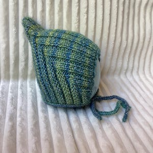 Könnte beinhalten: Eine handgestrickte Haube mit einem melierten Garnmuster in Blau- und Grüntönen. Die Haube hat eine spitz zulaufende Oberseite, eine durchgehende Rippenstruktur und zwei geflochtene Bänder, die an der Unterkante zum Binden dienen.