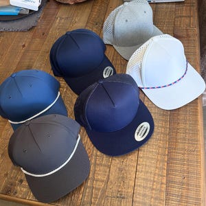 May include: Six baseball caps in shades of navy, grey, and white are displayed. Some have mesh backs, others are solid. Several feature a rope detail around the brim. The caps are arranged on a wooden surface. Measurements are not available.