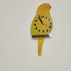 May include: A yellow parakeet-shaped wall clock with black numbers and hands. The clock has a red beak and black dots. The clock is mounted on a white wall.