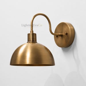 May include: A brass-colored wall-mounted light fixture with a curved arm and a dome-shaped shade. The fixture has a circular base and is mounted on a white wall. The text "Lighting Era Co" is visible on the fixture.