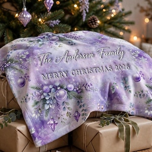 May include: A soft purple fleece blanket featuring a festive pattern of Christmas ornaments, pine branches, and snowflakes. The text "The Anderson Family" and "MERRY CHRISTMAS 2026" is printed in white across the center of the fabric.