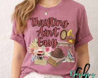 May include: A mauve-colored t-shirt with the phrase "Thrifting Ain't Easy" in a decorative font. The graphic includes vintage items like a gramophone, hat, teacup, and books. The shirt has rolled-up sleeves.