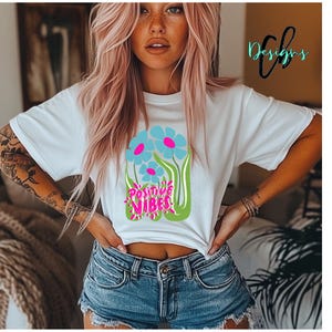 May include: White t-shirt with a floral design featuring blue flowers with pink centers and the words "Positive Vibes" in pink and green. The shirt has short sleeves.