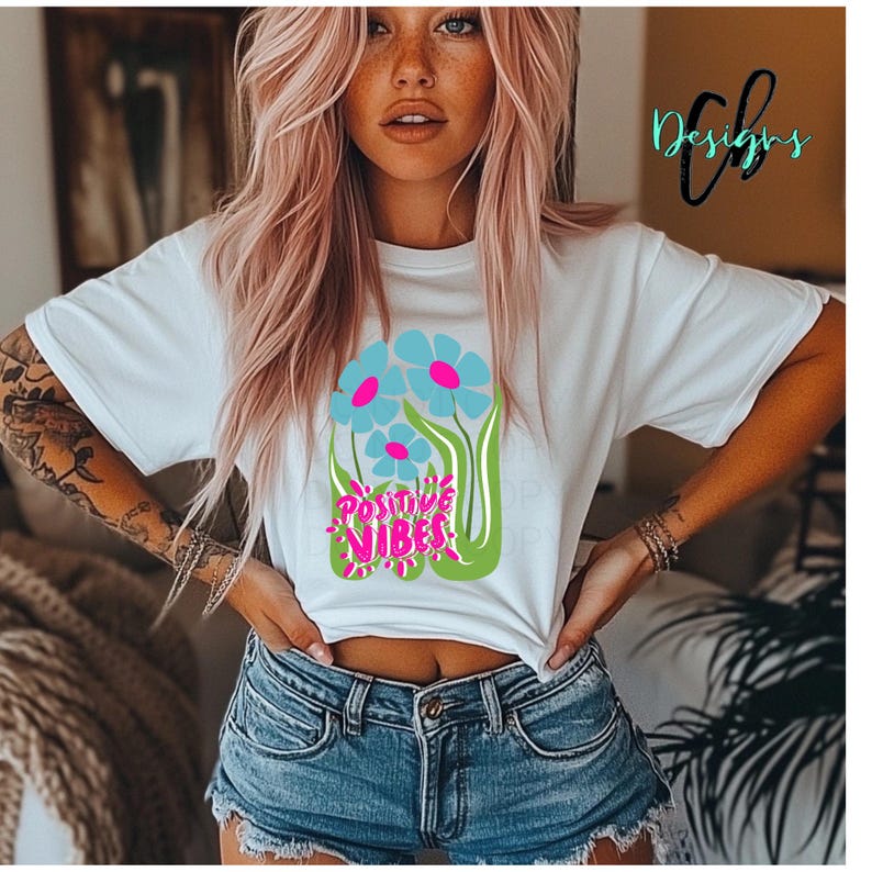 May include: White t-shirt with a floral design featuring blue flowers with pink centers and the words "Positive Vibes" in pink and green. The shirt has short sleeves.