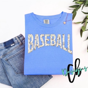 May include: A light blue t-shirt with the word "BASEBALL" in a camouflage pattern. The shirt is paired with blue jeans. A gold pendant necklace is draped on the shirt. Green leaves are in the background.