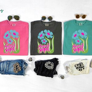 May include: Three t-shirts in pink, charcoal, and teal, each with a floral design and the words "Positive Vibes." The image also includes denim shorts, sunglasses, and sneakers.