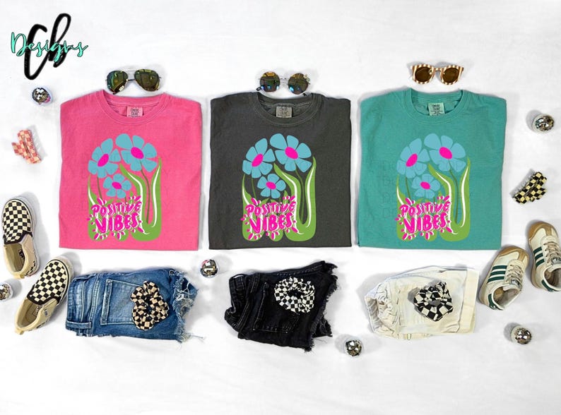 May include: Three t-shirts in pink, charcoal, and teal, each with a floral design and the words "Positive Vibes." The image also includes denim shorts, sunglasses, and sneakers.