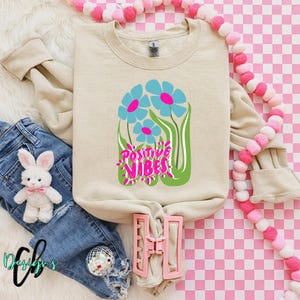 May include: A beige sweatshirt with a floral design featuring blue and pink flowers and the words "Positive Vibes" in pink and green. The sweatshirt is paired with blue jeans and a plush bunny.