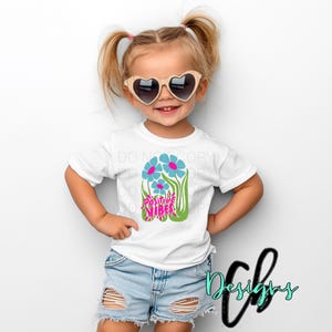 May include: A young child wearing a white t-shirt with a floral design and the words "Positive Vibes". The shirt is paired with denim shorts and heart-shaped sunglasses. The child is smiling.