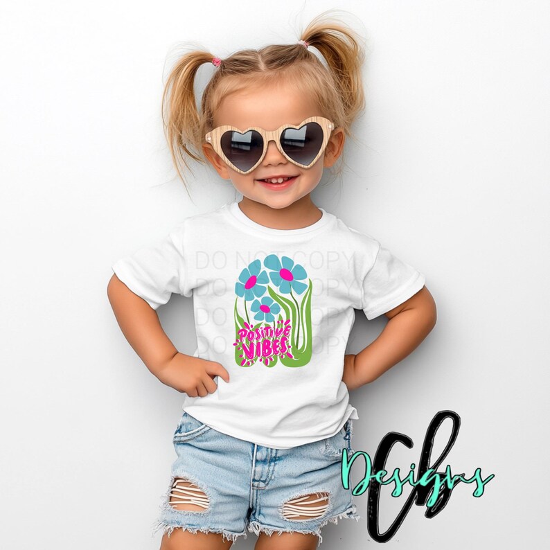 May include: A young child wearing a white t-shirt with a floral design and the words "Positive Vibes". The shirt is paired with denim shorts and heart-shaped sunglasses. The child is smiling.