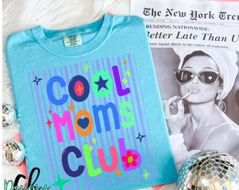 May include: A light blue t-shirt with the words "COOL MOMS CLUB" in colorful letters. The design includes stars, a heart, and a flower. A newspaper clipping and a disco ball are in the background.