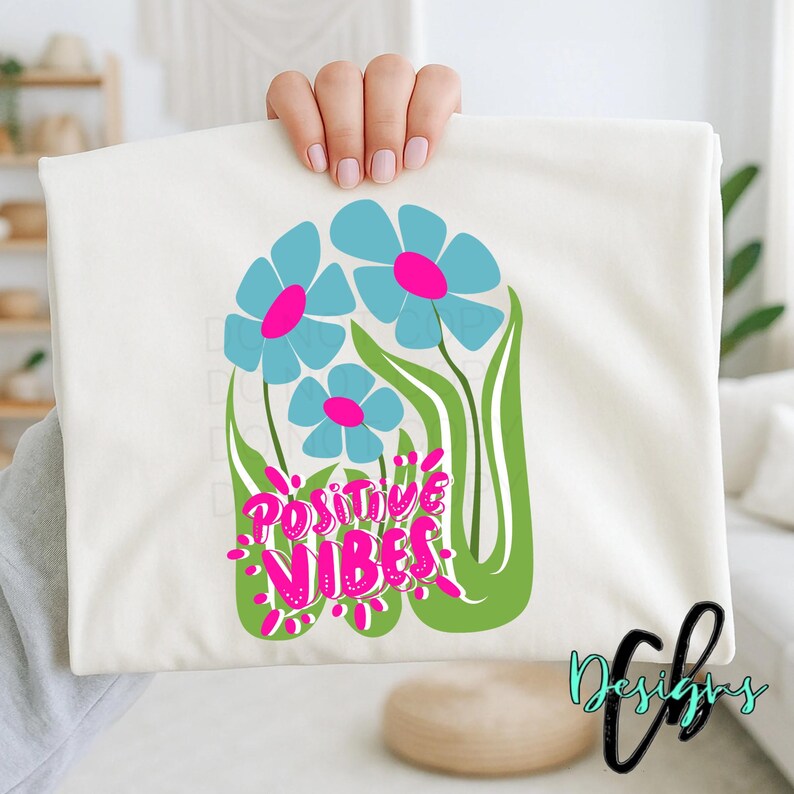 May include: Cream-colored t-shirt featuring a floral design with three blue flowers with pink centers and green stems. The words "Positive Vibes" are written in pink and green script. The shirt is held up by a person.